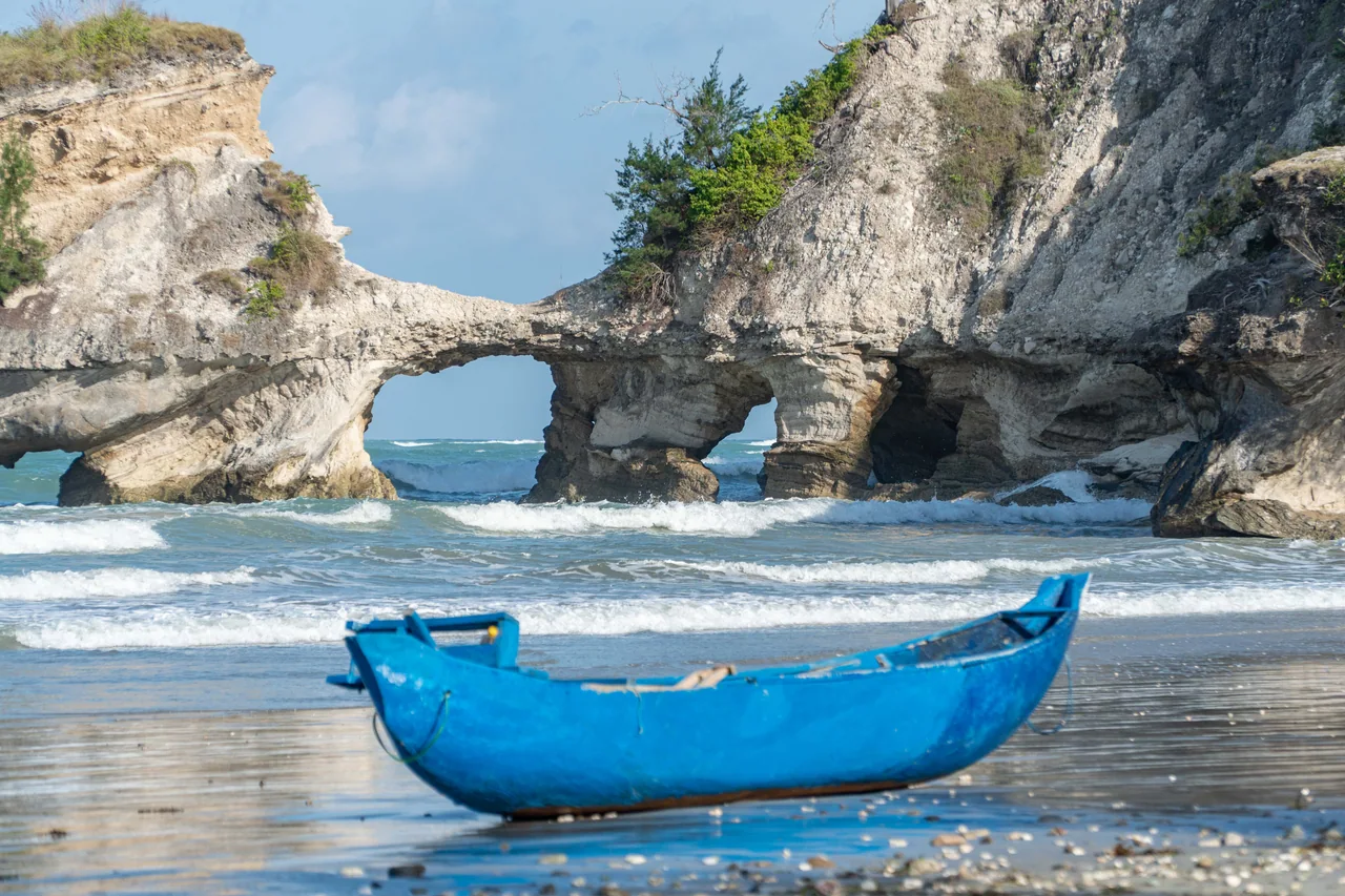 A blue boat on the sand beneath the limestone rock opening at Watu Parunu